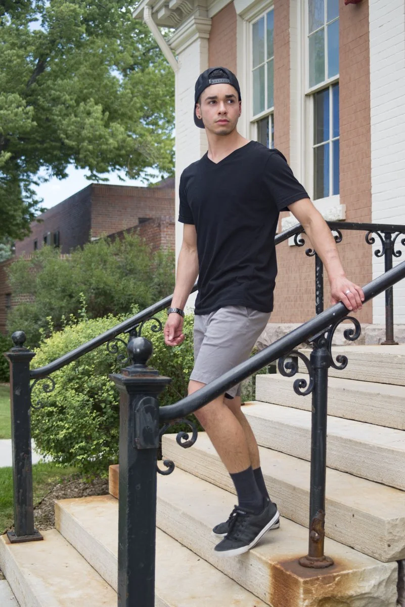 A young man with a black cap, black T-shirt, gray shorts, and black sneakers is walking down concrete stairs outside a brick house with large windows. There are trees and greenery around.