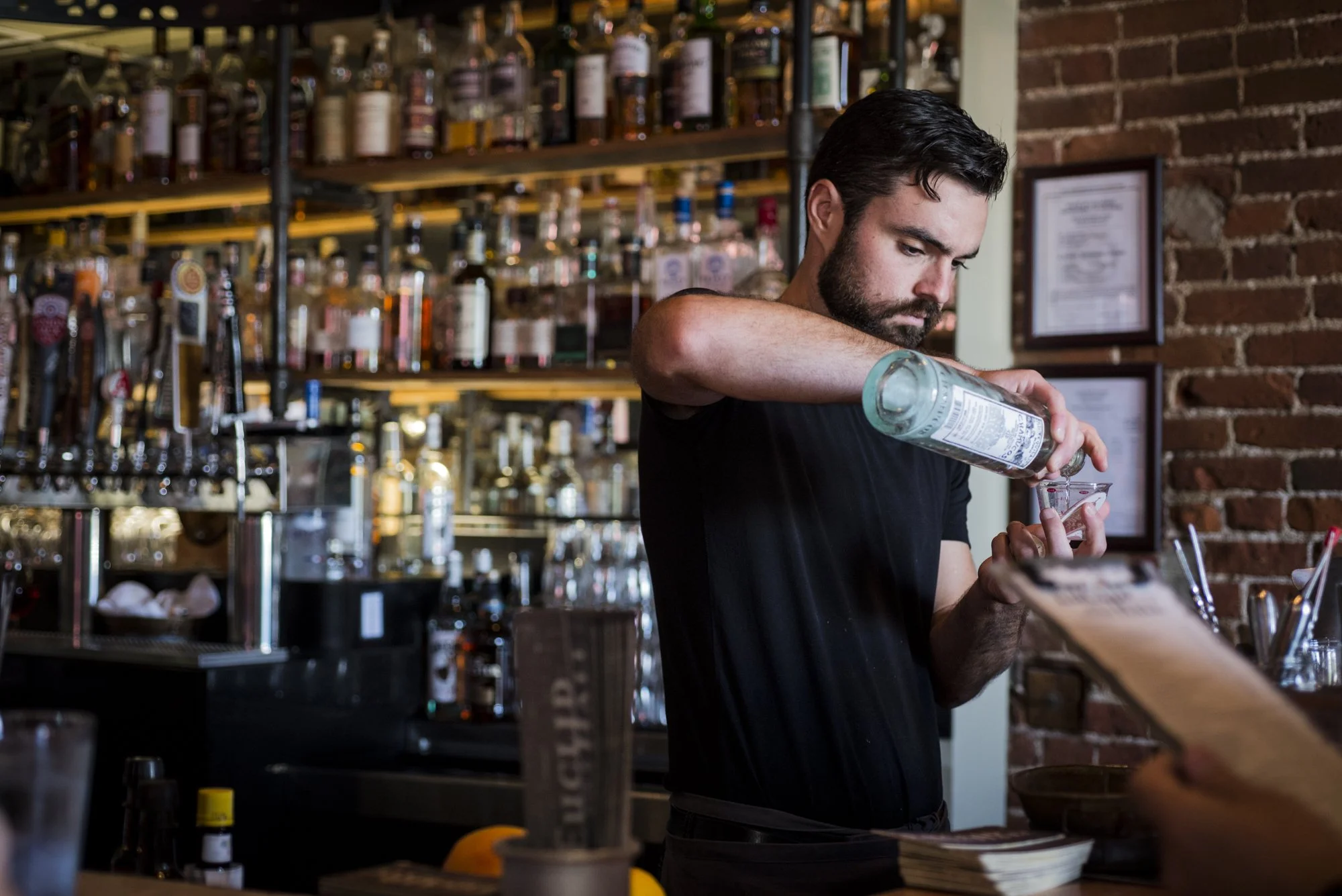 Man with dark hair and beard pouring a clear liquid into a glass in a bar with an industrial style, exposed brick wall, and shelves filled with various liquor bottles.