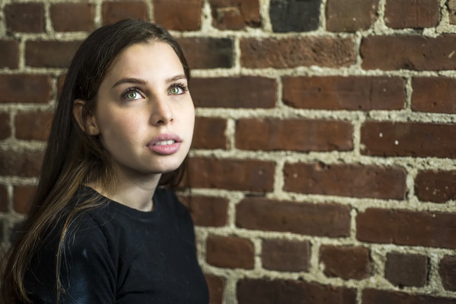 A young woman with long brown hair and green eyes, wearing a black shirt, standing against a red brick wall and looking slightly upward.