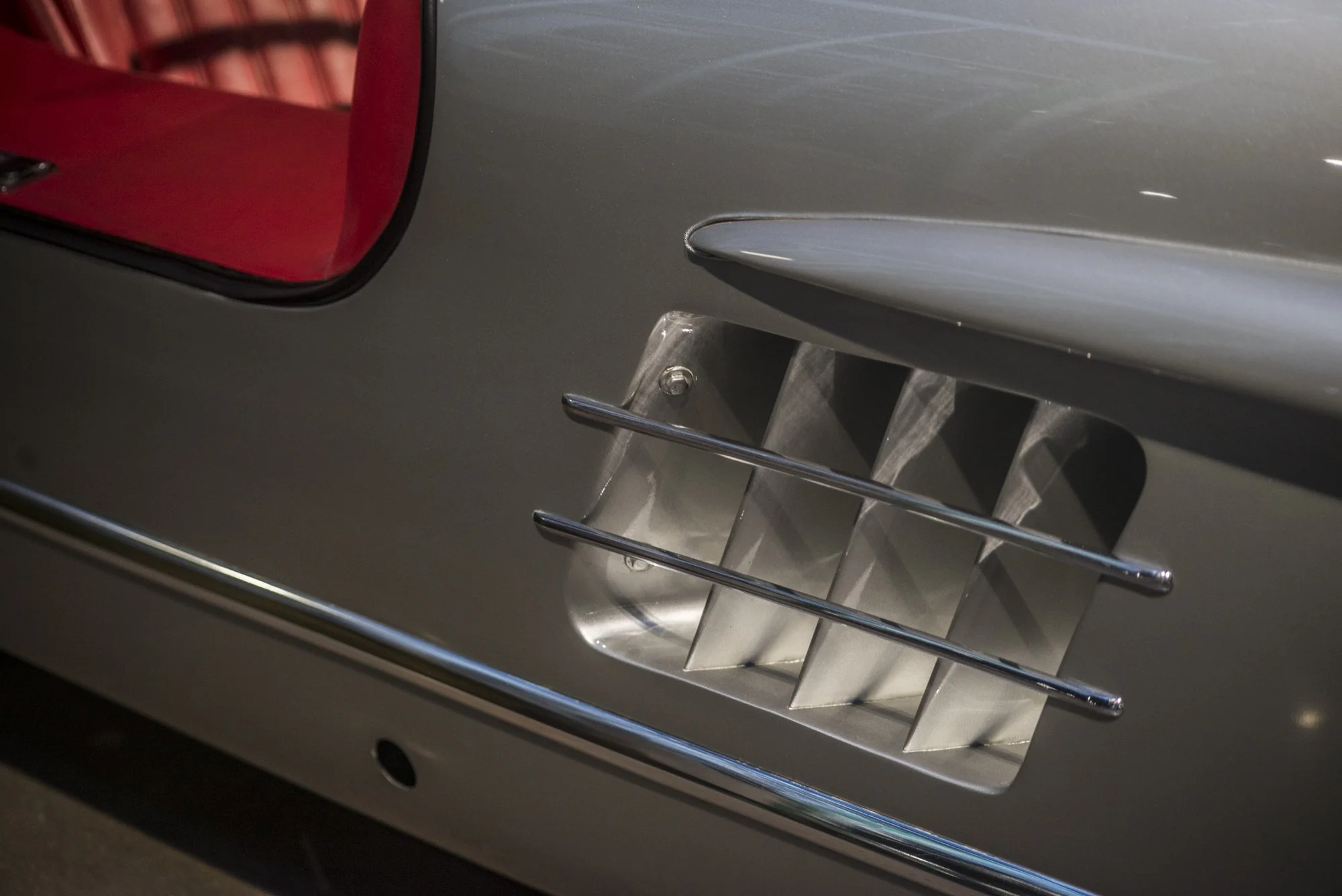 Close-up image of the front part of a classic silver car, including a grille with horizontal bars and a part of the headlight and red interior seat.