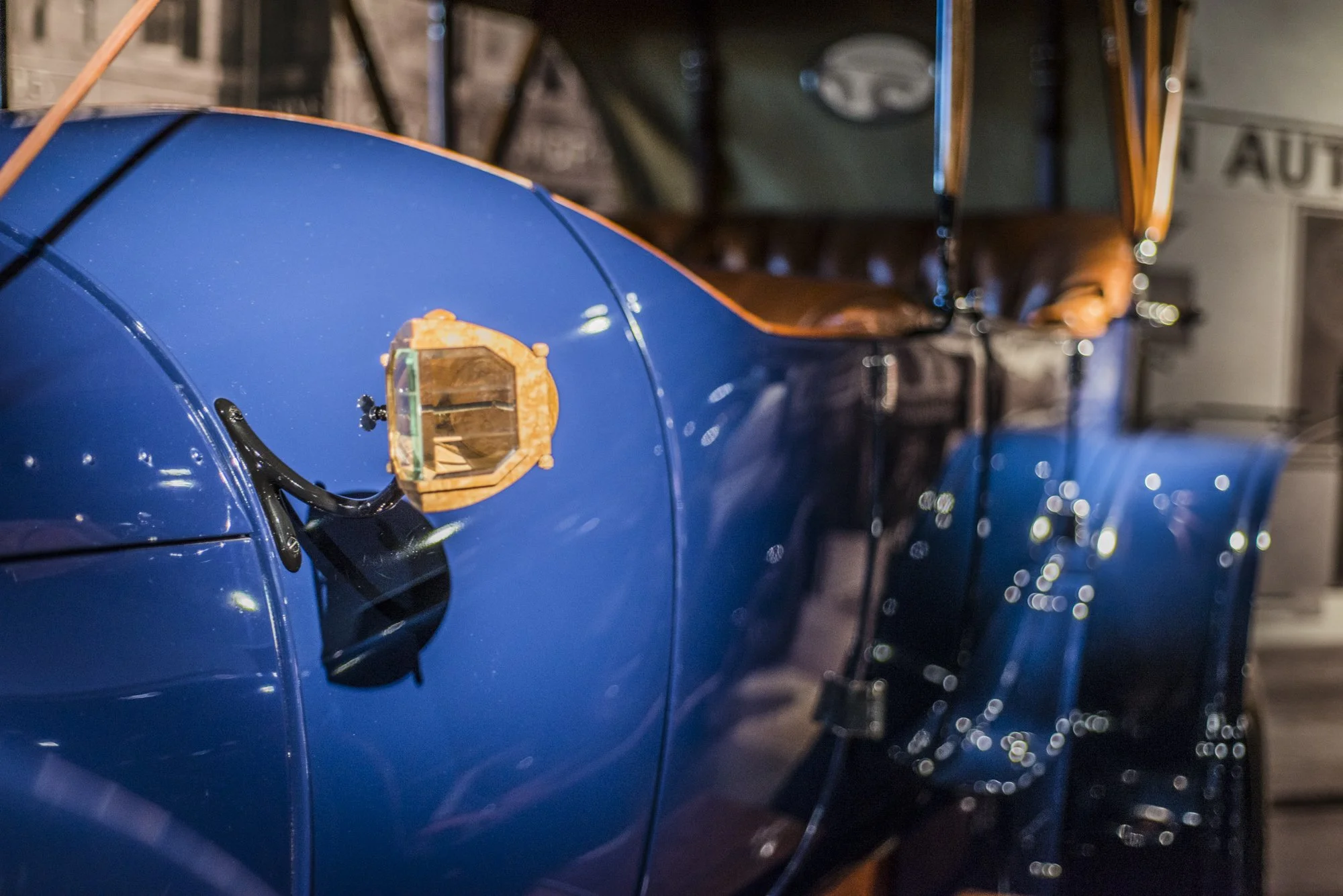 A close-up view of a vintage blue automobile with a brown leather interior and a small mirror on the side. Blurred background with lights.