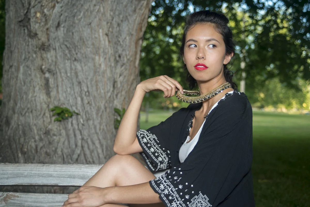 A young woman with dark hair and red lipstick, wearing a black embroidered cover-up, sitting on a wooden bench outdoors near a large tree with green foliage in the background, holding a layered gold necklace near her neck.
