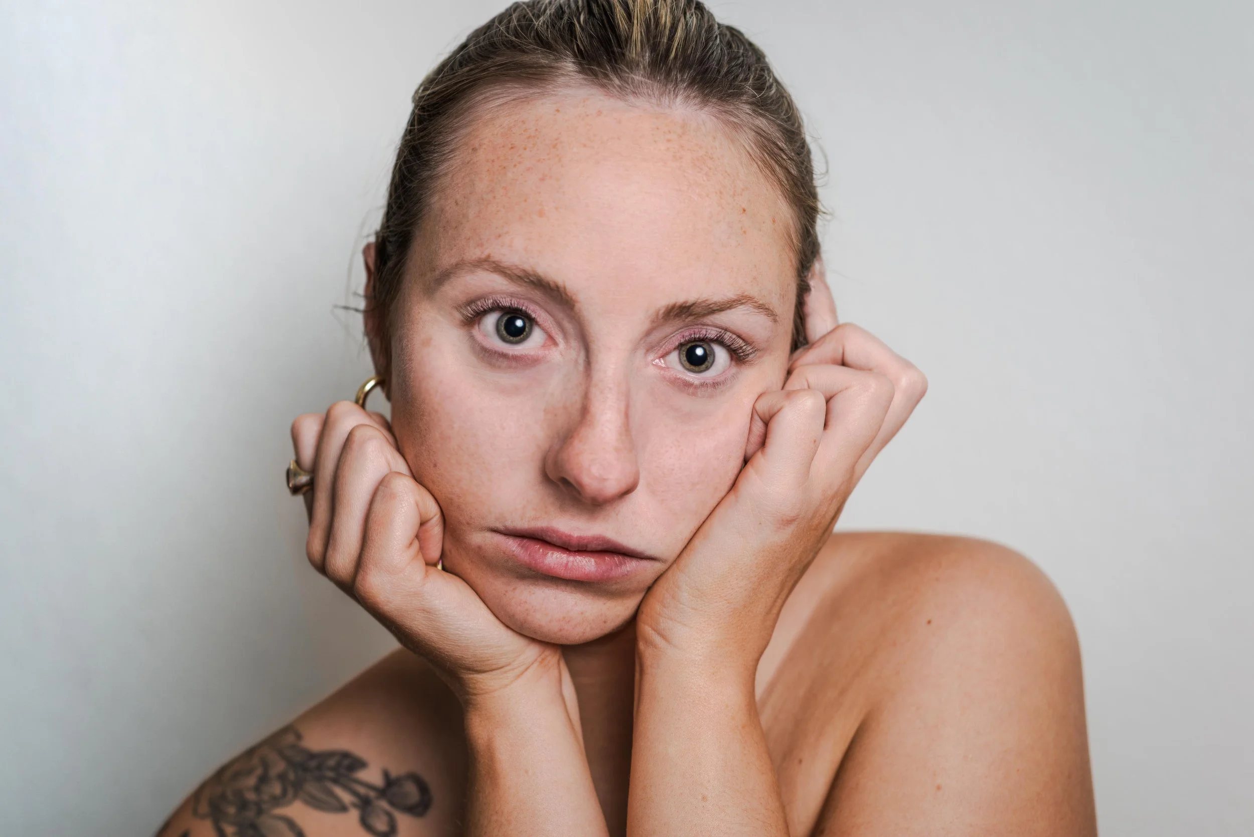A woman with light skin and freckles looks directly at the camera. She has her hands on her cheeks, with her fingers slightly pressing her face. She has light brown hair pulled back and is wearing gold earrings. The background is plain and light-colo