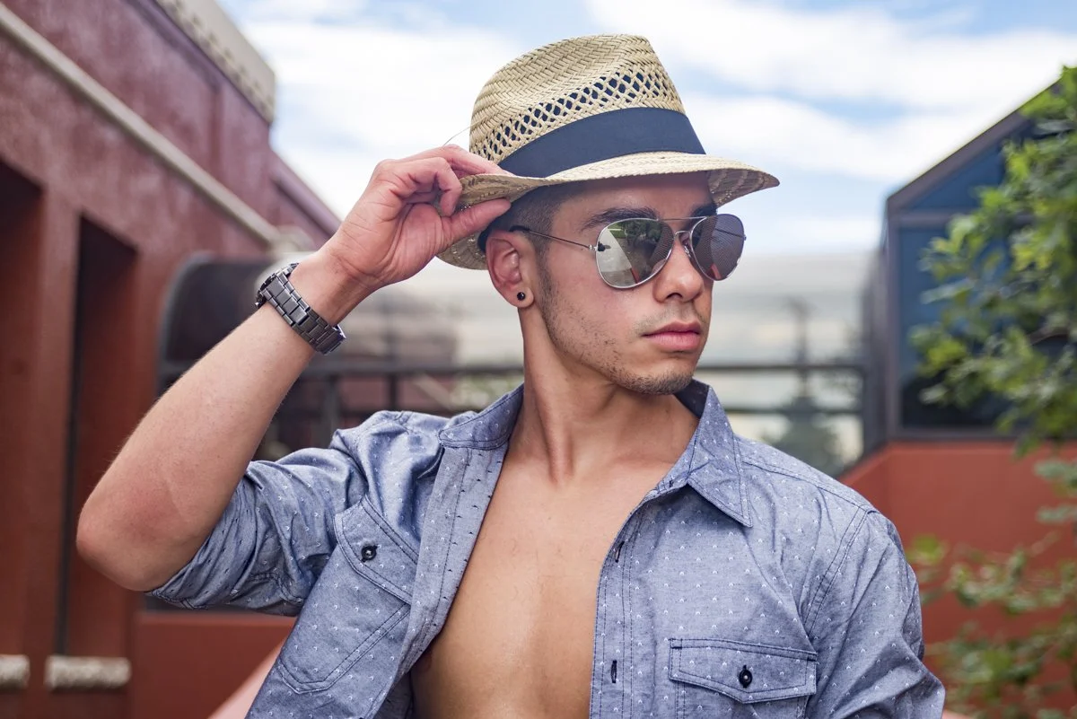 A young man wearing sunglasses and a straw hat, partially unbuttoned blue shirt, standing outdoors, with colorful buildings and greenery in the background.