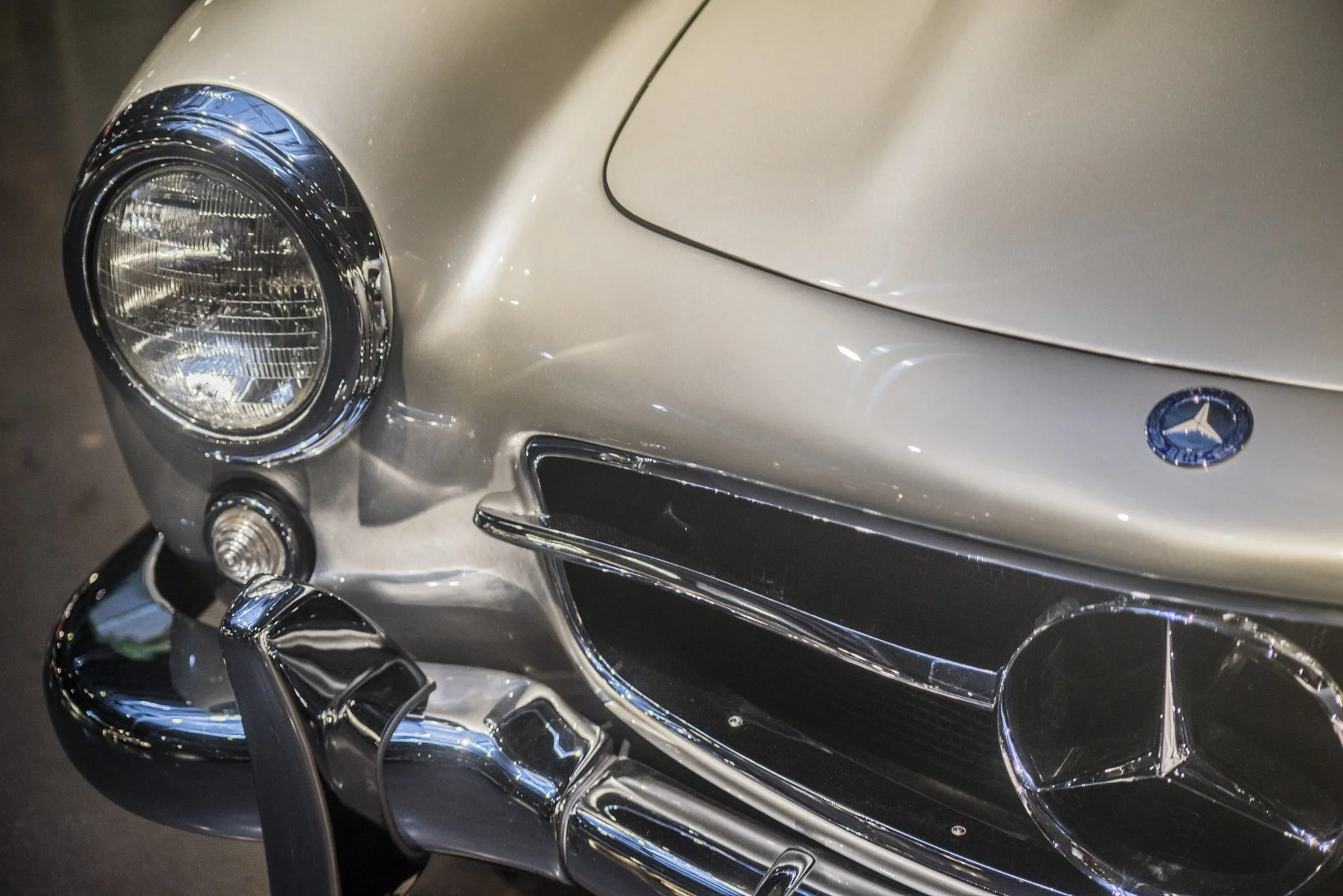 Close-up of a silver Mercedes-Benz classic car, showing the front left headlight, part of the grille, and the Mercedes-Benz logo on the hood.