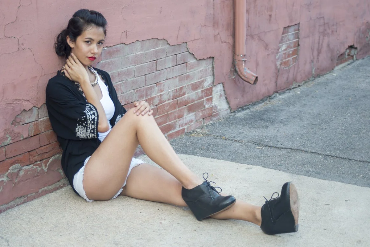 A young woman sits on the sidewalk against a pink, weathered brick wall. She wears a black embroidered jacket, white shorts, and black ankle boots, with a necklace and red lipstick. Her hair is styled in loose curls.