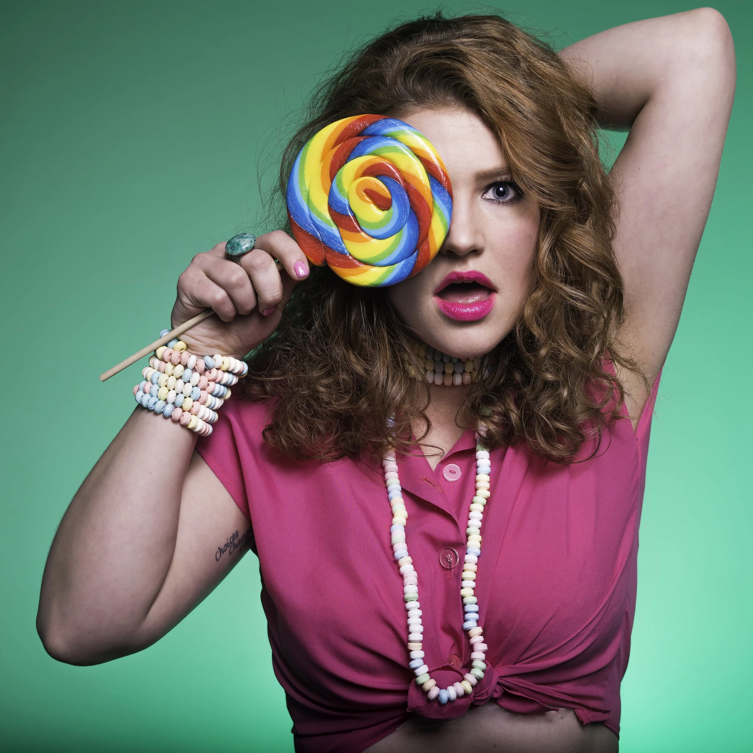 Woman with curly red hair wearing a bright pink polo shirt, holding a rainbow-colored lollipop over her right eye, against a solid teal background.