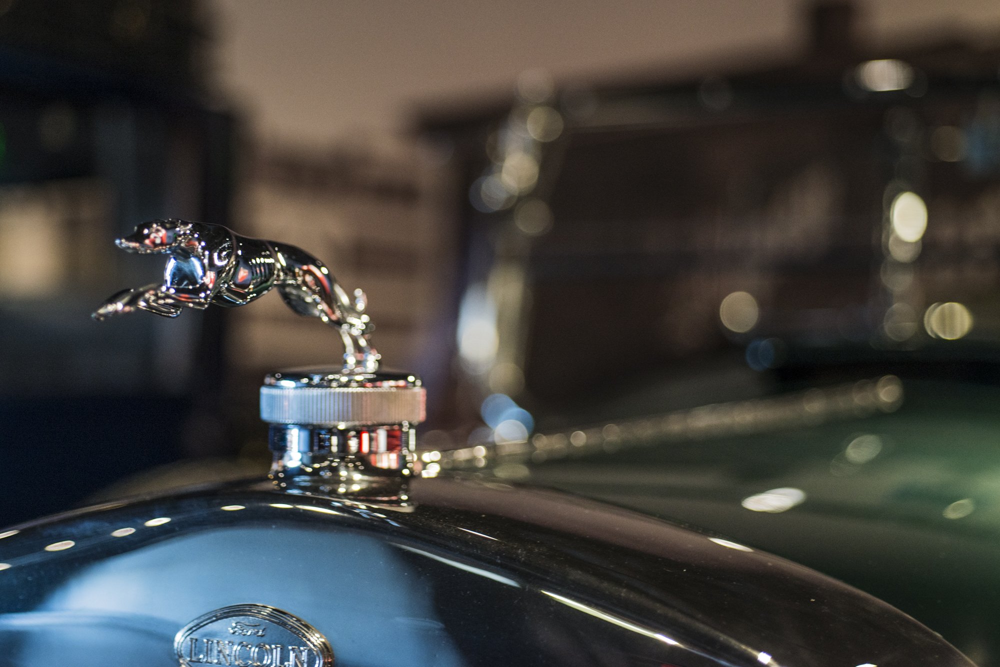 Close-up of a silver Lincoln hood ornament on a black vehicle, with blurred background of garage or workshop.