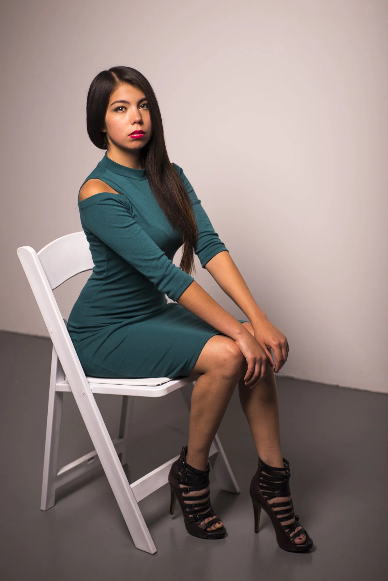 A woman with long dark hair, wearing a teal dress with cutouts on the shoulders, sitting on a white chair. She has red lipstick and is wearing high-heeled black and brown strappy shoes. The background is plain and neutral.
