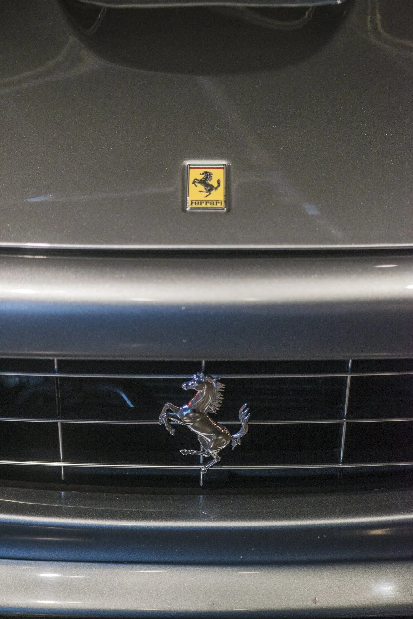 Close-up of a black Ferrari hood ornament with the Ferrari emblem, and a Ferrari grille below with a silver prancing horse badge.