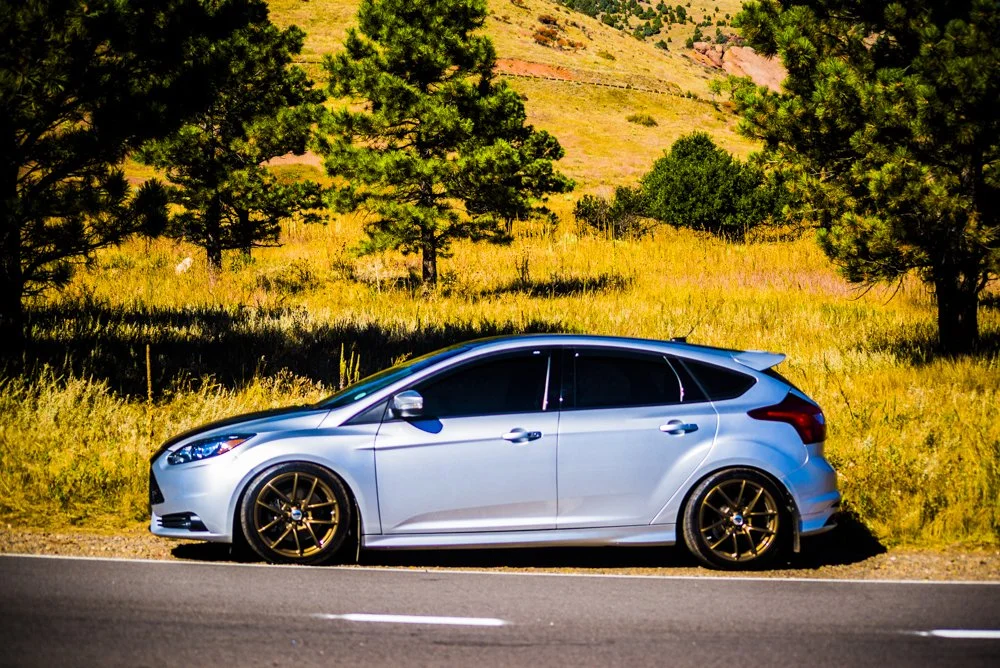 A silver hatchback car with black wheels parked on the side of a road, with a grassy hillside and trees in the background.