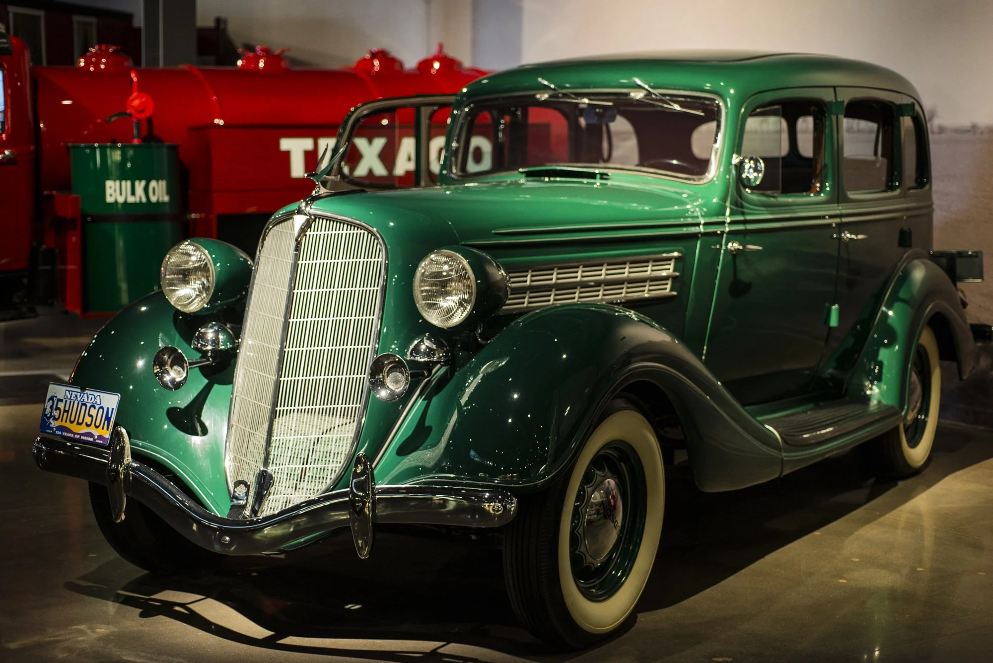 A vintage green car with white-walled tires on display indoors. Behind it, there is a red oil tank labeled 'BULK OIL' and a vintage red petrol pump with a sign reading 'TAXA'.