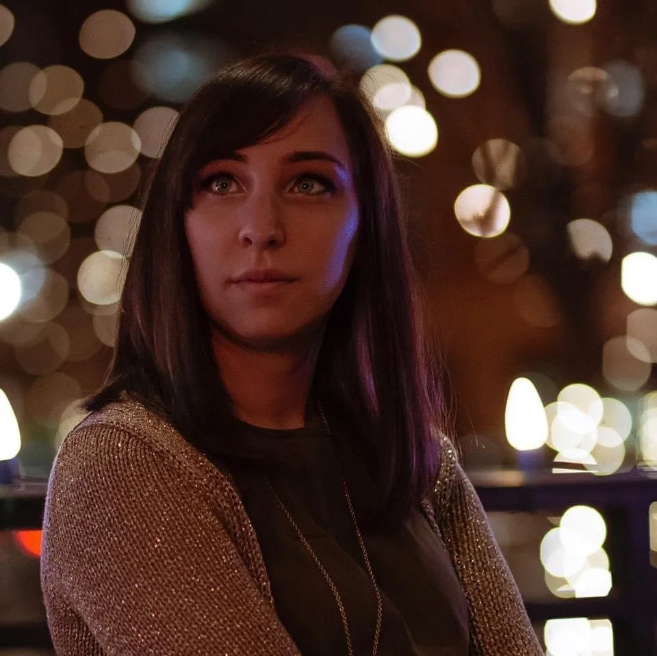 A woman with shoulder-length dark hair and light skin looking to her right, illuminated by colorful lights in the background.