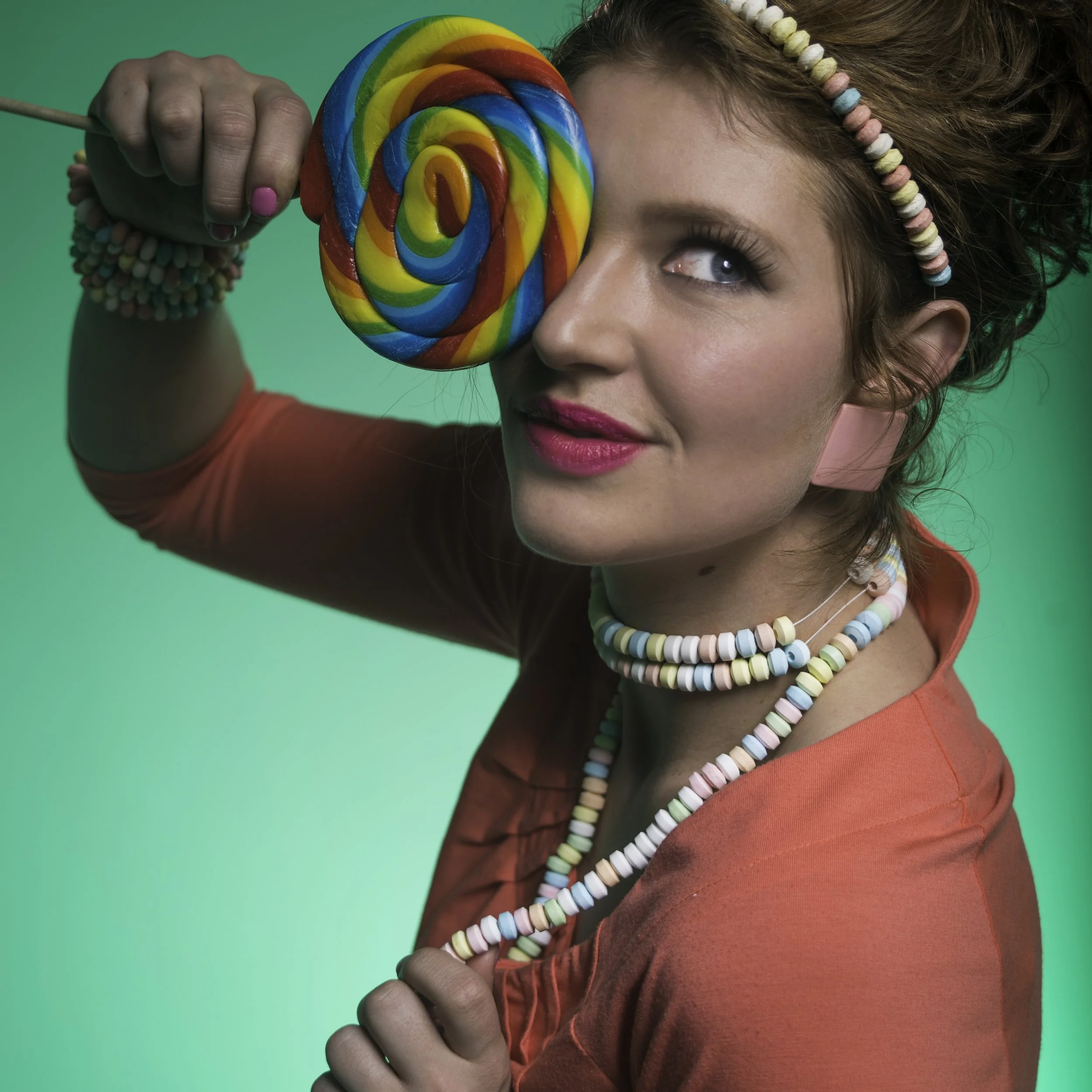 A woman with curly hair holding a large rainbow-colored lollipop in front of her face, with colorful beaded necklaces, a beaded headband, and pink lipstick, against a green background.