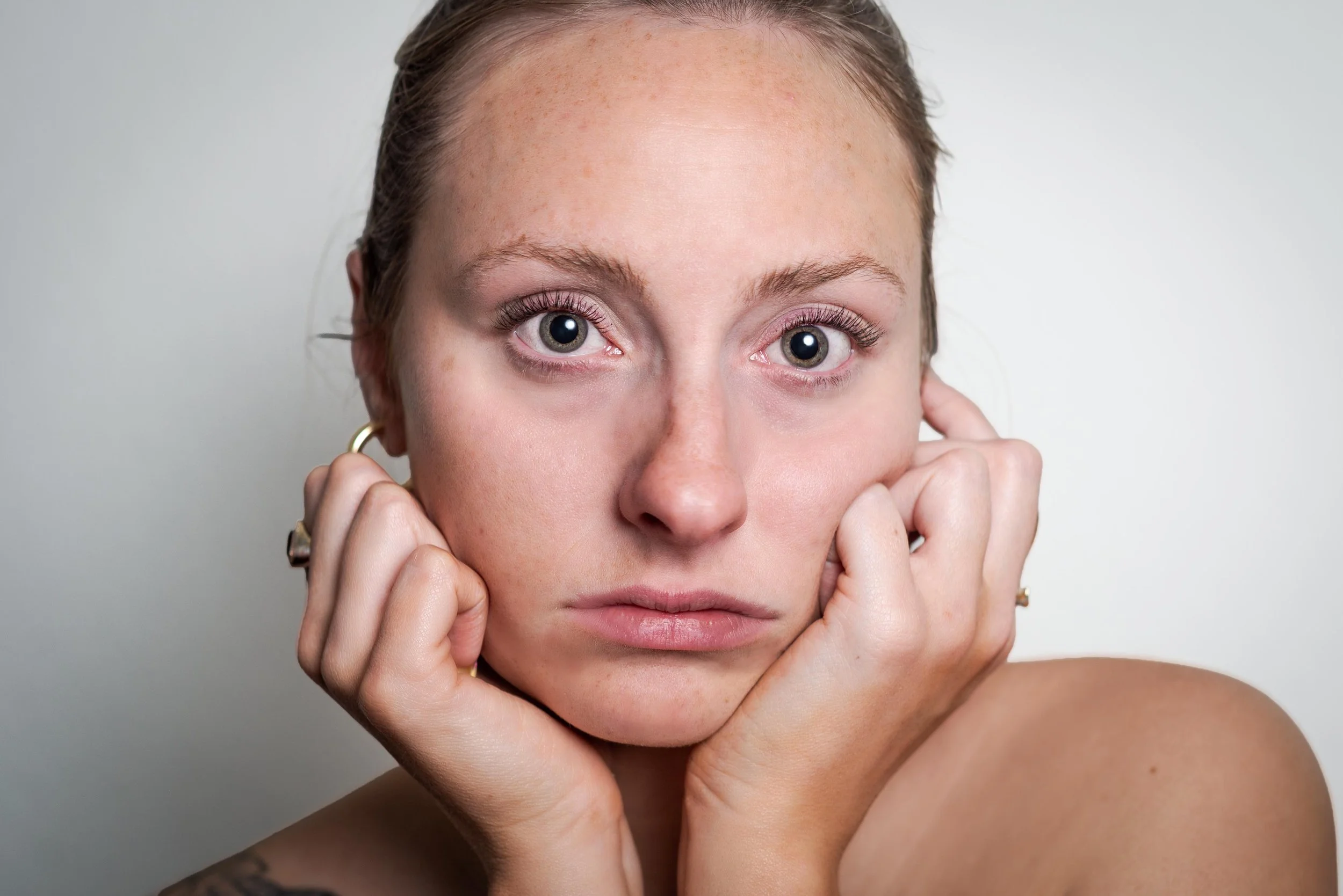 Close-up portrait of a woman with blue eyes, freckles, and light brown hair pulled back, holding her face with both hands against a neutral background.