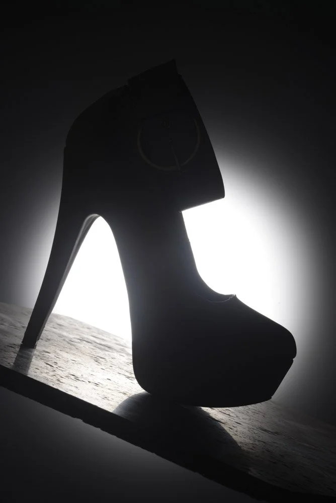 Silhouette of a high-heeled shoe with a platform, viewed against a backlit background on a wooden surface.