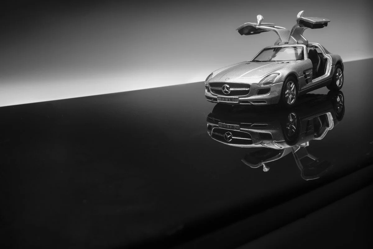 A black and white photo of a model Mercedes-Benz car with its gullwing doors open, placed on a reflective surface.