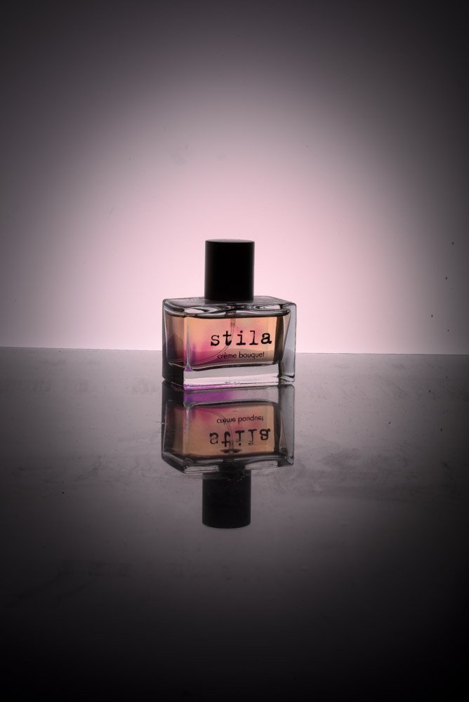 A bottle of Stila Creme Bouquet perfume with a black cap, reflected on a shiny surface against a soft pink and gray gradient background.