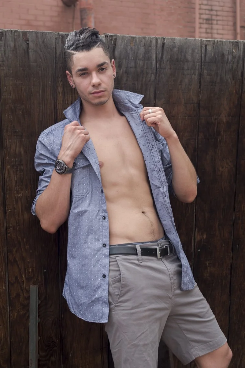 A young man with dark hair styled upward, wearing a partially open blue button-up shirt and khaki shorts, standing against a wooden fence outside.