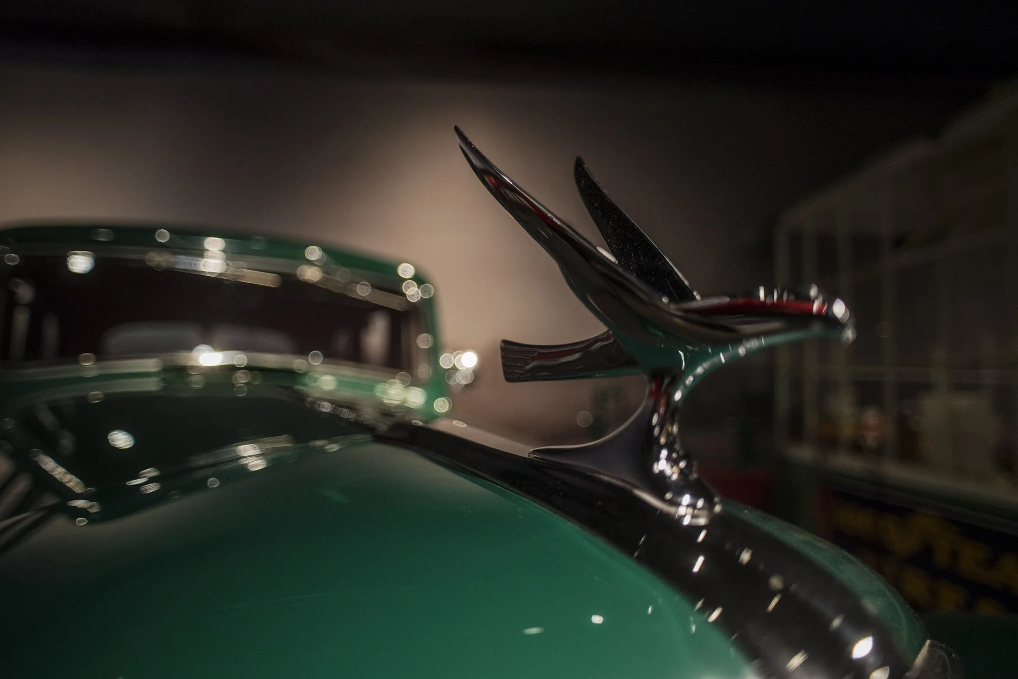 Close-up of the front of a vintage green car with chrome detailing and a prominent chrome hood ornament, blurred background and reflection of lights.