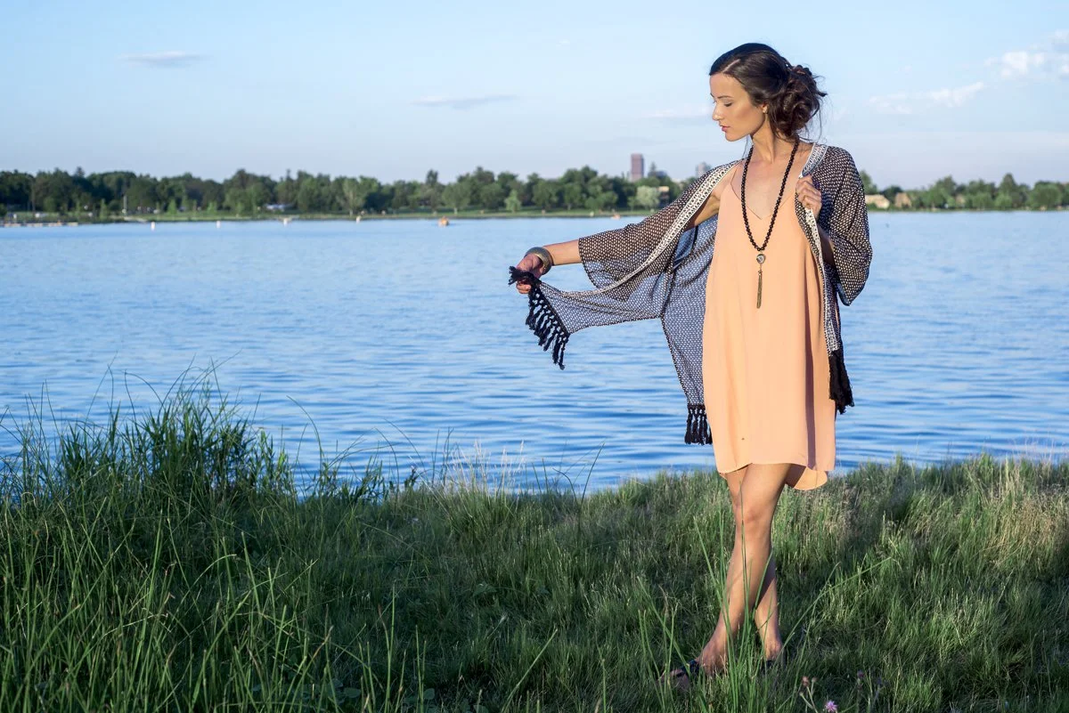 A woman standing on the grassy edge of a lake, wearing a peach dress and a black and white shawl, holding the shawl with one hand and looking down thoughtfully, with trees and a cityscape in the background under a partly cloudy sky.