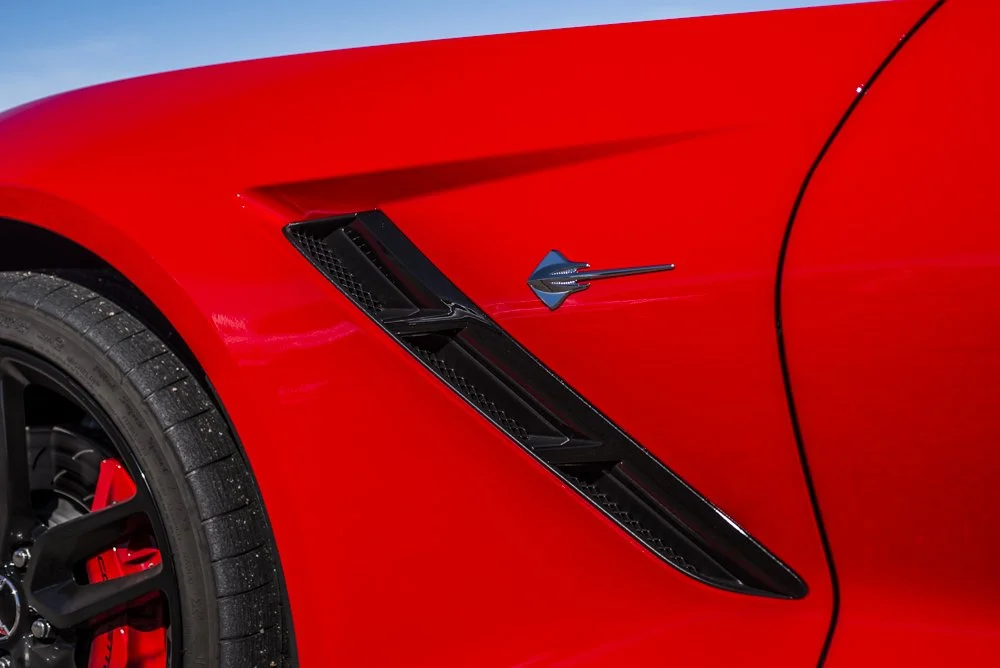 Close-up of a red sports car's front fender, featuring a black air vent and a blue arrow-shaped emblem.