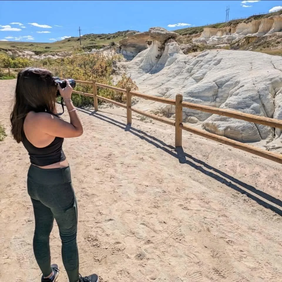 A woman in athletic clothing takes a photo with binoculars in a desert-like area with rocky terrain and a wooden fence.