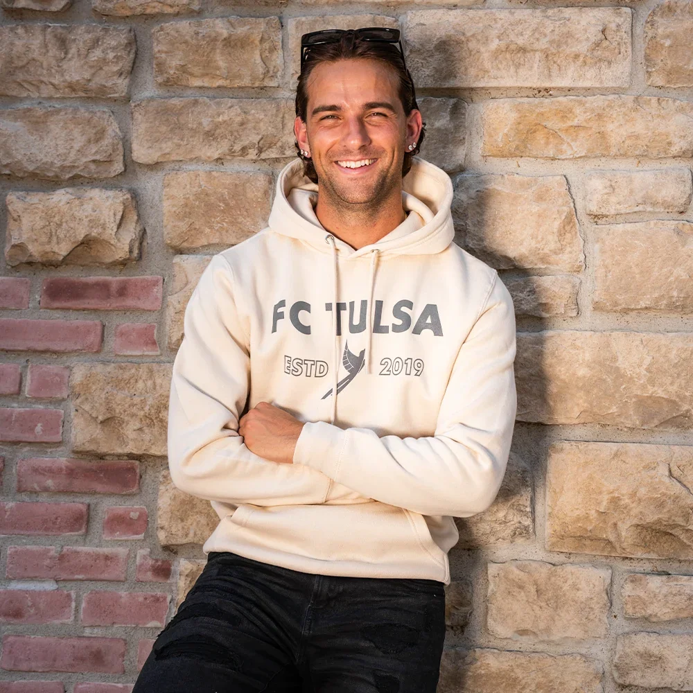 A smiling man with sunglasses on his head, earrings, and a beige hoodie with 'FC TULSA' on it, leaning against a stone wall.