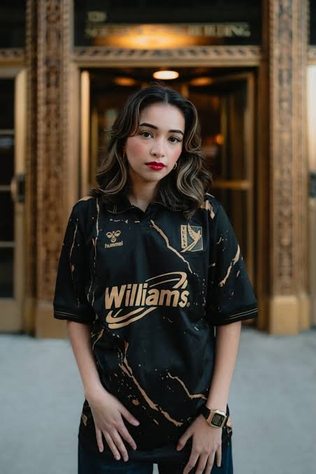 Young woman standing in front of a decorative background, wearing a black sports jersey with gold accents and the word "Williams" on it.