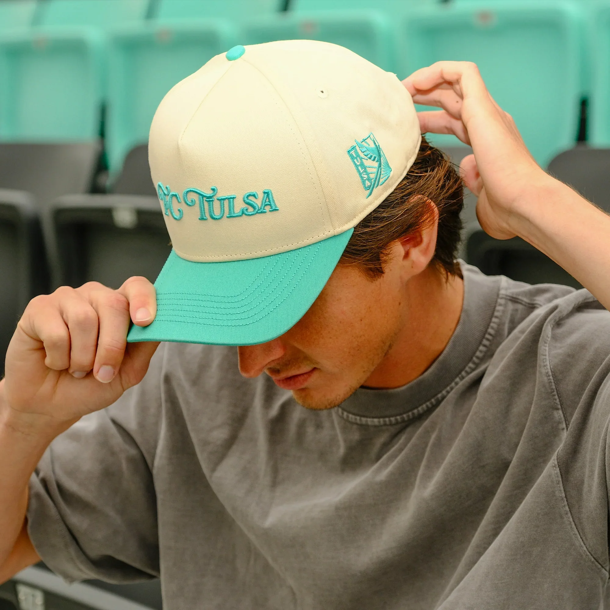 A person wearing a baseball cap with the words HC TULSA embroidered on it, adjusting the cap with one hand while wearing a gray hoodie.