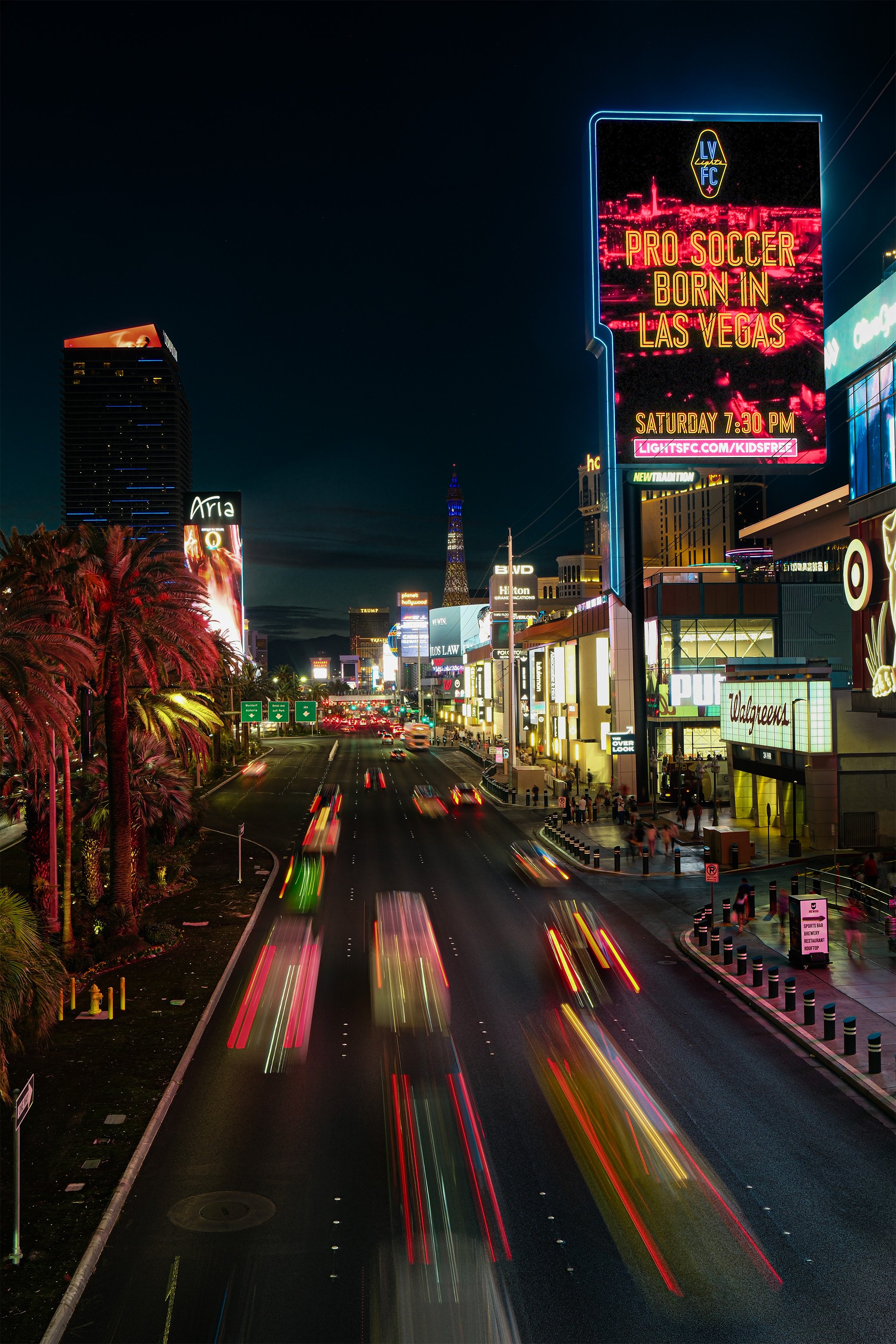 Night view of Las Vegas Boulevard with light trails from moving cars, illuminated billboards including one promoting pro soccer, palm trees on the left, and a skyline with buildings and neon signs.