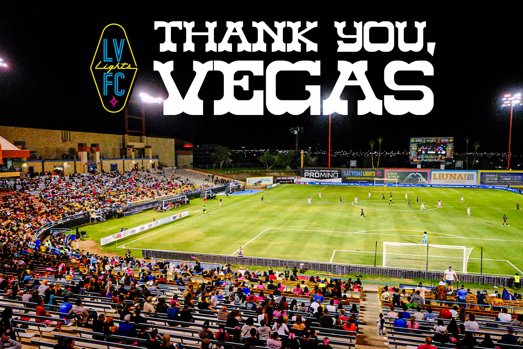 A nighttime soccer game at a stadium with a large crowd of spectators. The field is lit by bright stadium lights and shows players in action. The background features advertising banners and a large screen, with the words "Thank You, Vegas" and the logo "LV Lights FC" in neon-style text.