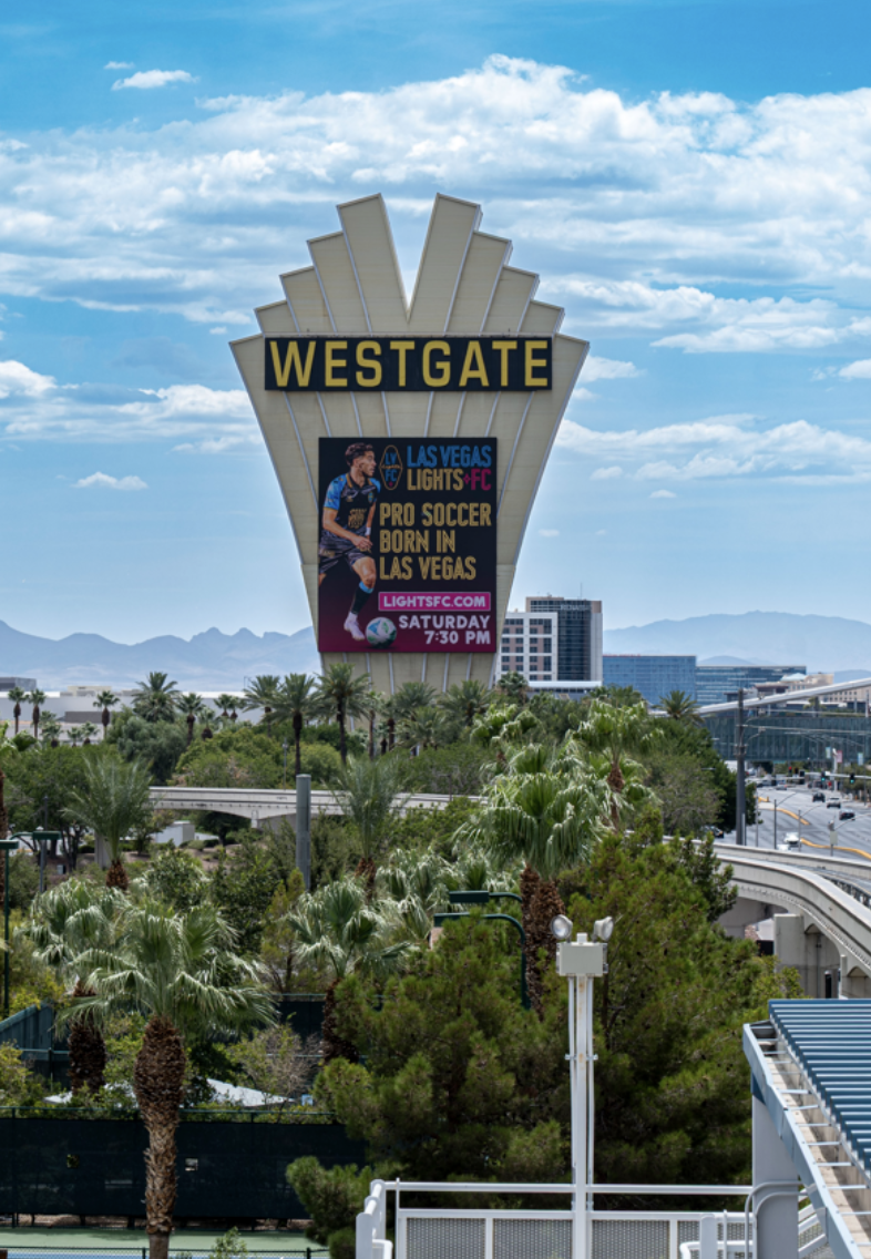 Large sign for Westgate with a colorful advertisement for a pro soccer event in Las Vegas, featuring a player and event details, set against a blue sky with clouds and a cityscape with trees in the foreground.