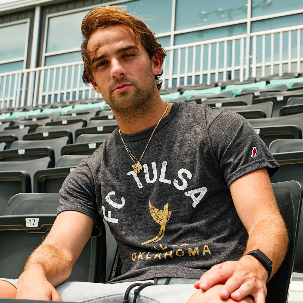 A young man with brown hair, wearing a gray T-shirt with 'FC Tulsa Oklahoma' and a gold-colored logo, sitting in an empty stadium with black seats.