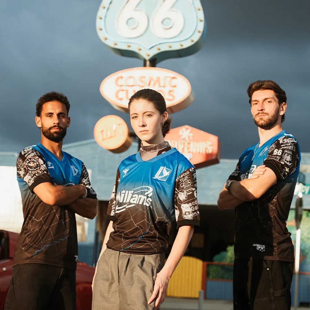 Three young adults, two men and one woman, standing outside in front of a retro-style amusement park sign at dusk. They are wearing matching blue and black racing jerseys with various logos.