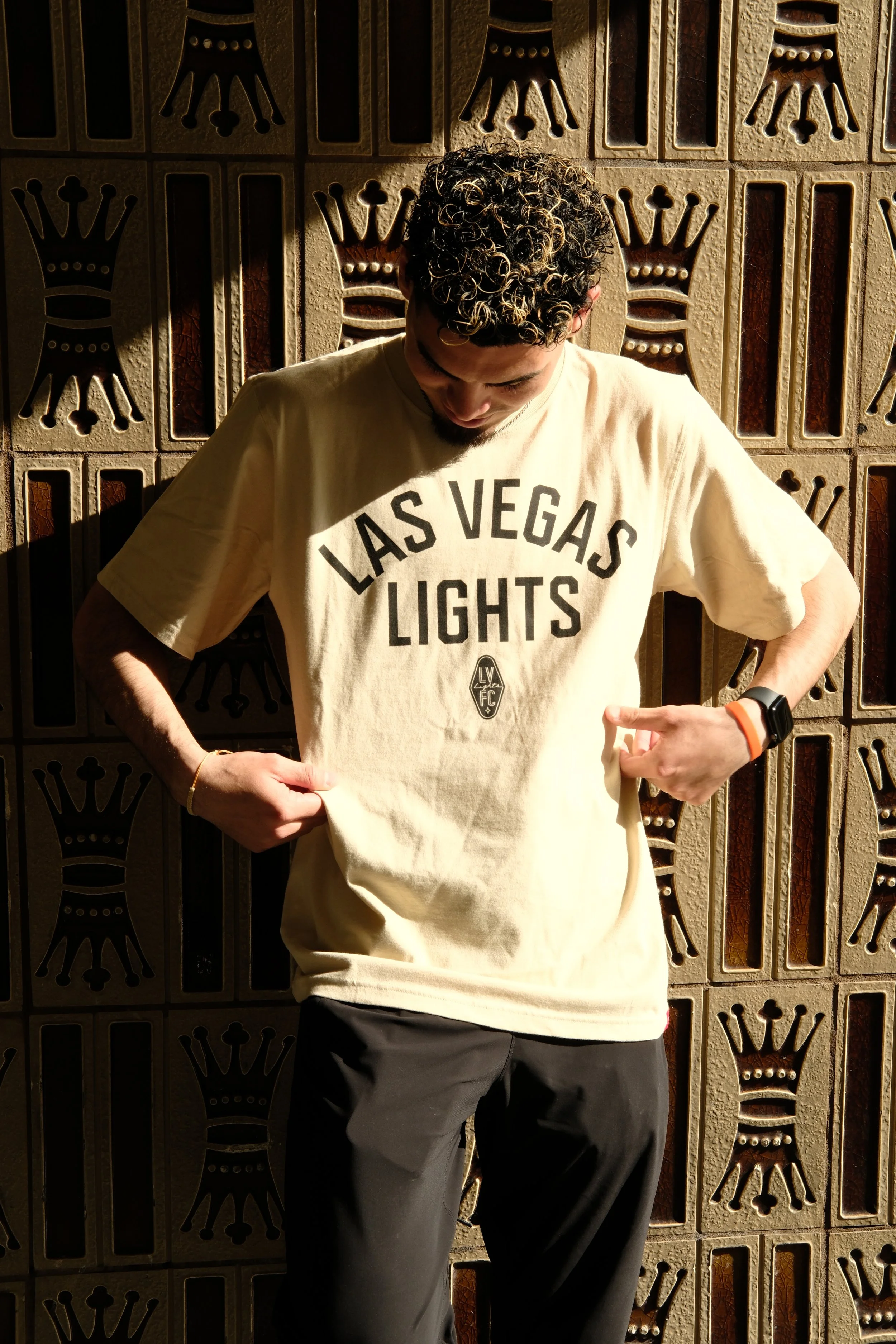 A young man with curly hair wearing a beige T-shirt with the words 'Las Vegas Lights' and a logo, standing in front of a decorative metal wall with crowns and arches, looking down and pointing to his shirt with his right hand.