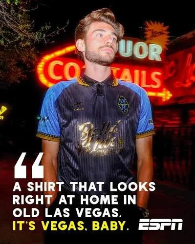 A young man with styled brown hair and a beard wearing a dark blue shirt with gold and blue accents standing in front of neon signs at night in Las Vegas. He has a confident expression, and the image includes overlays with the ESPN logo and the quote, "A shirt that looks right at home in old Las Vegas. It's Vegas, baby."