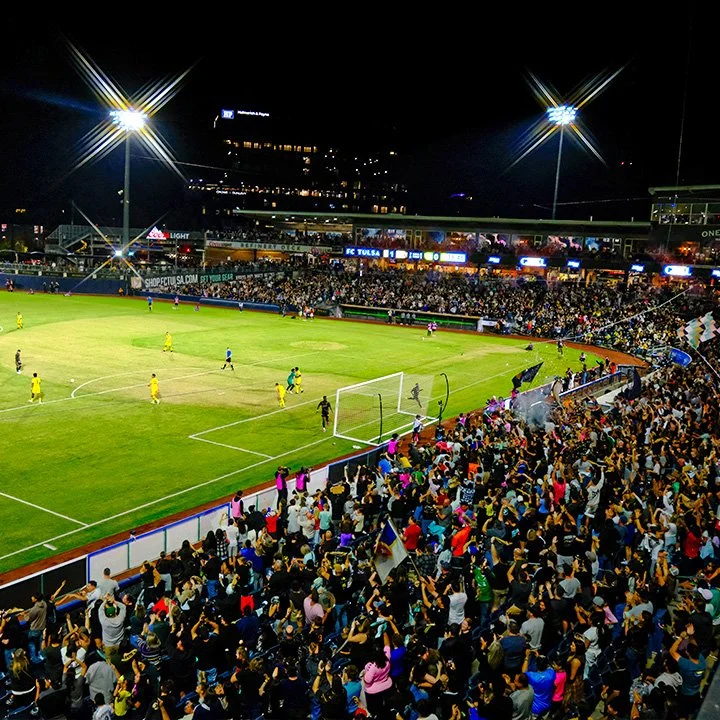 Nighttime soccer match in a large stadium filled with cheering fans, with players on the field, some near the goal, and bright stadium lights overhead.