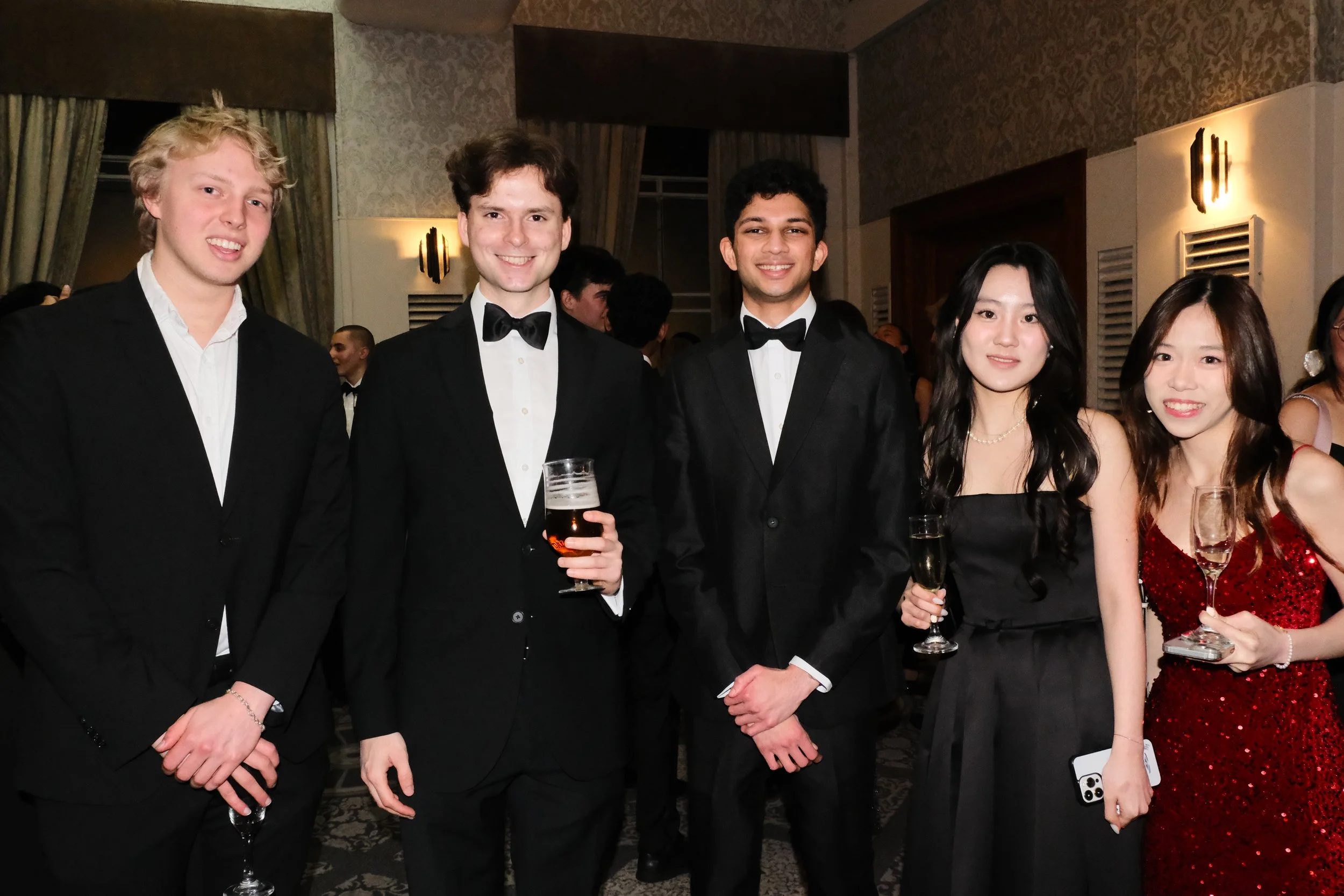 Group of five young adults dressed in formal attire, smiling at a social event in a decorated room with patterned wallpaper, curtains, and wall sconces. The men are wearing tuxedos, and the women are in elegant dresses, holding drinks and a phone.