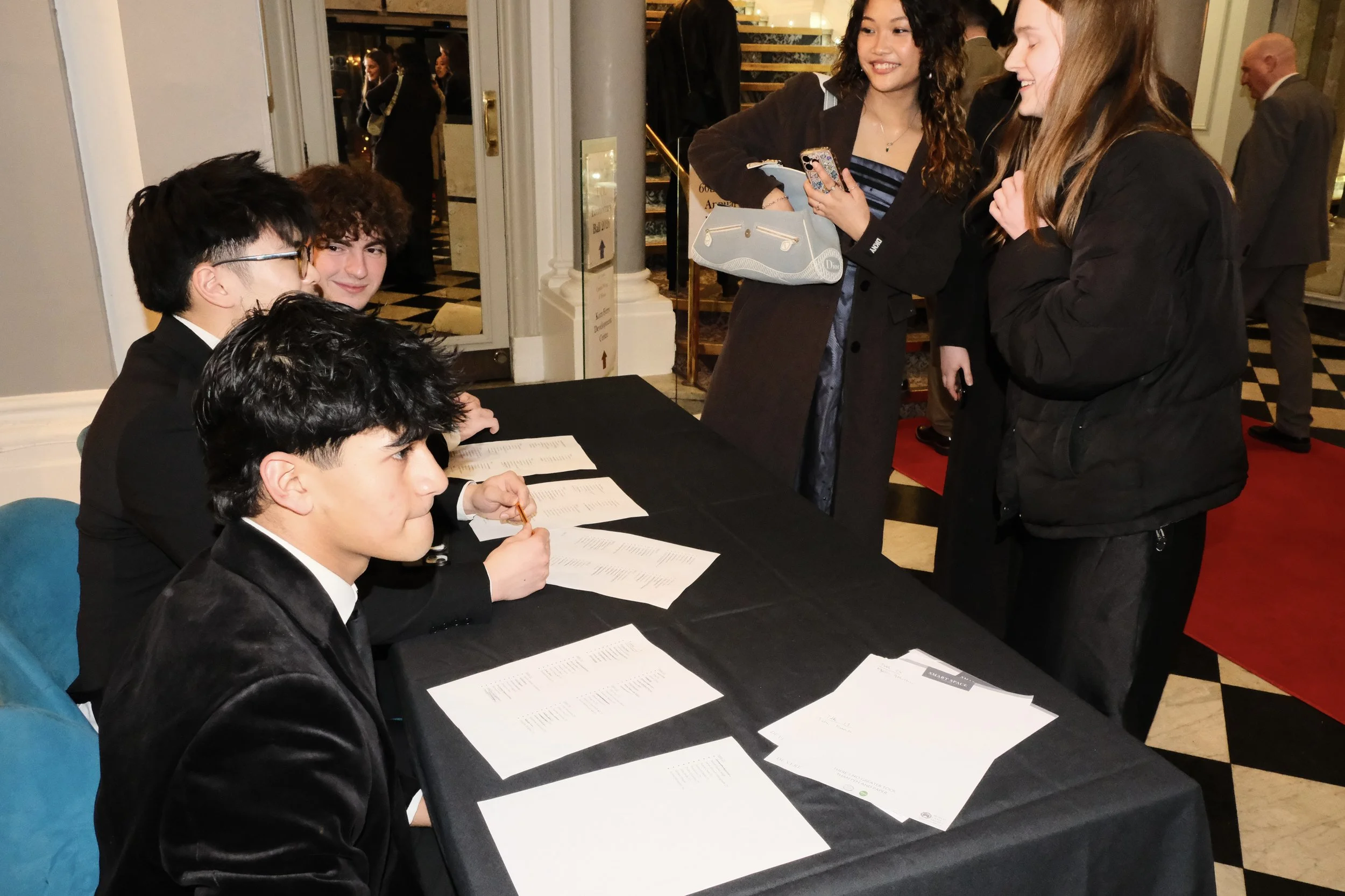 People gathered around a registration or check-in table at an event, smiling and engaging in conversation. The table has papers and documents, and the setting appears to be an elegant indoor venue.