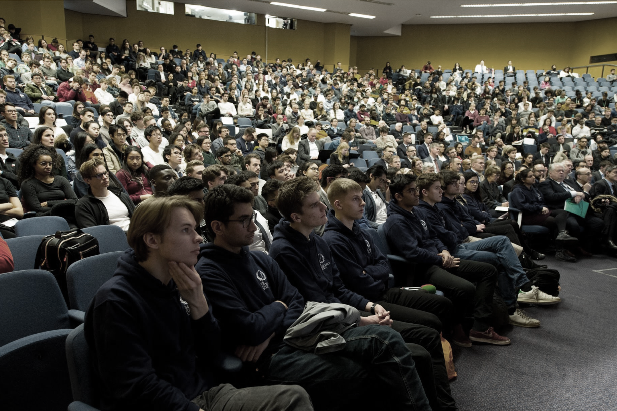 A large audience of diverse students and adults seated in a lecture hall or auditorium, attentively listening to a presentation or speaker.