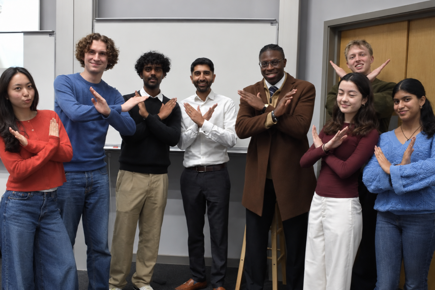Group of nine diverse young adults standing in front of a whiteboard, making X and O hand gestures and smiling.