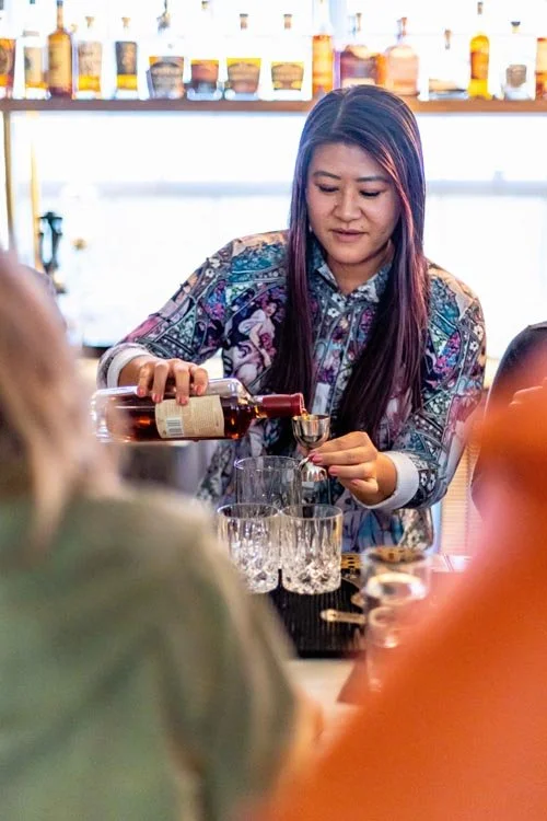 A woman pouring a brown liquor into a glass at a bar, with multiple glasses and a colorful background of bottles.