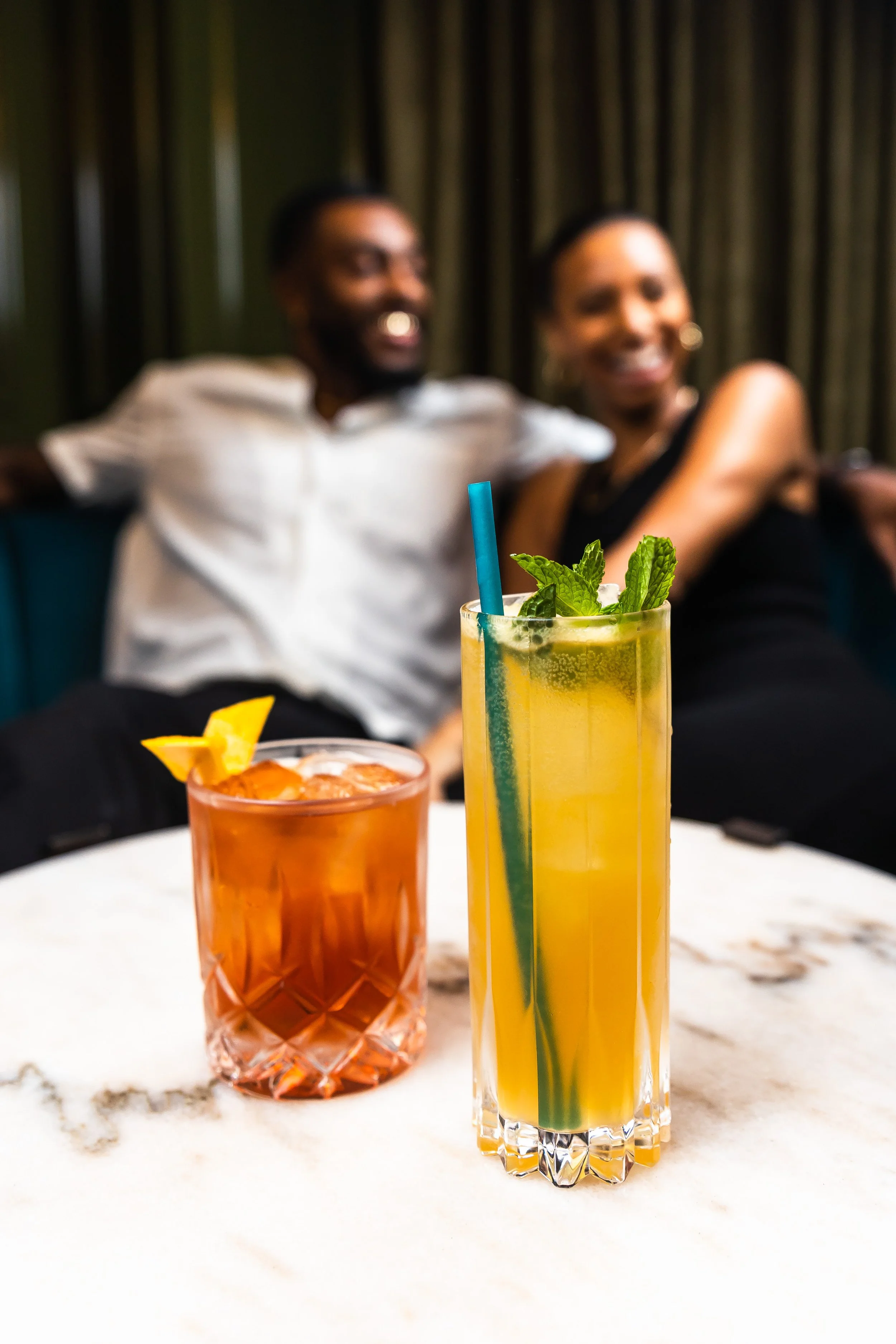 Two colorful cocktails on a marble table, with a blurred background of a happy couple smiling.