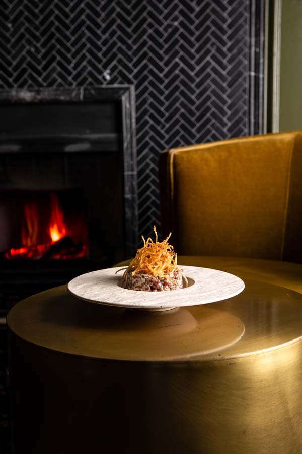 A plate with a gourmet dish garnished with crispy fried noodles served on a marble riser, placed on a round, reflective gold table next to a yellow velvet chair, with a black fireplace and dark patterned wall in the background.