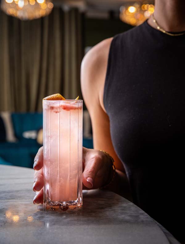 A woman holding a tall glass of pink cocktail garnished with citrus peel, in an upscale bar or restaurant setting.