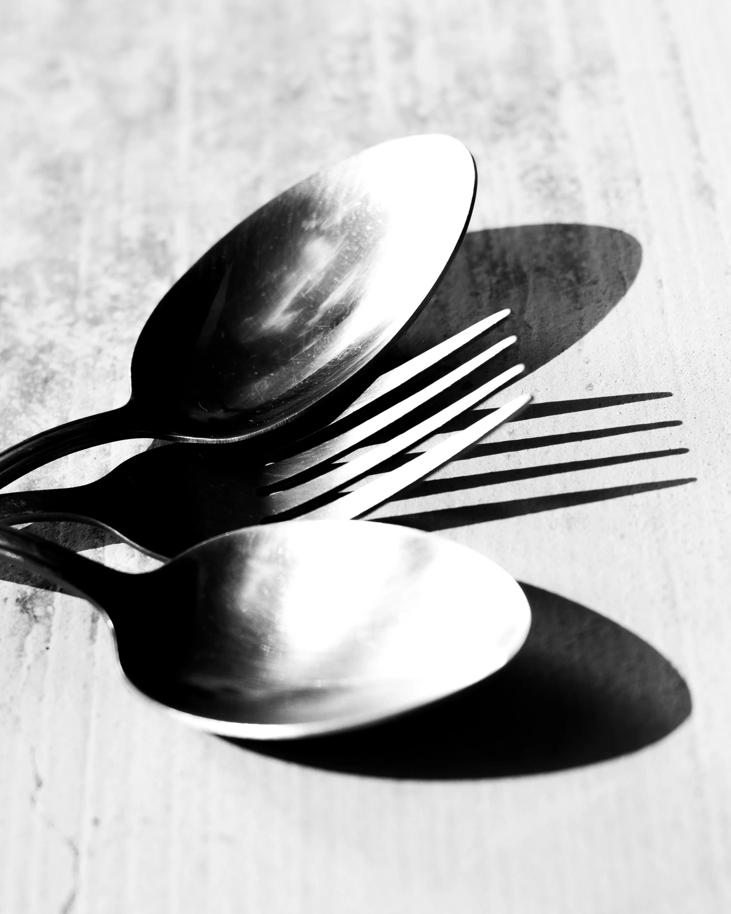 Close-up of a metal spoon and fork on a light-colored surface with shadows cast by the utensils.