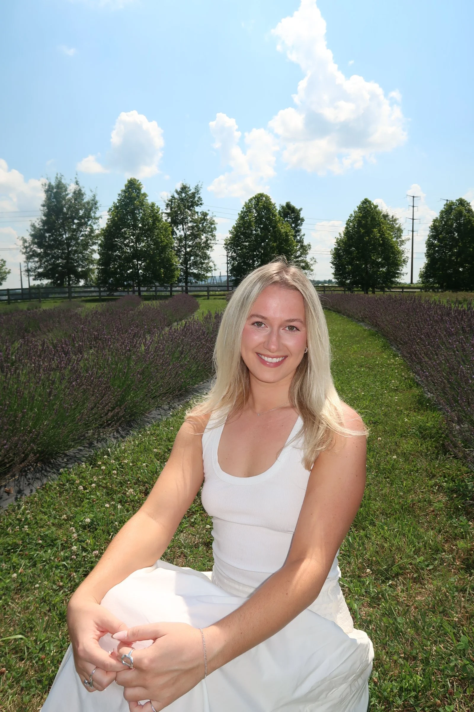 A woman with long blonde hair sitting outdoors in a grassy field surrounded by lavender plants and green trees, under a partly cloudy sky.