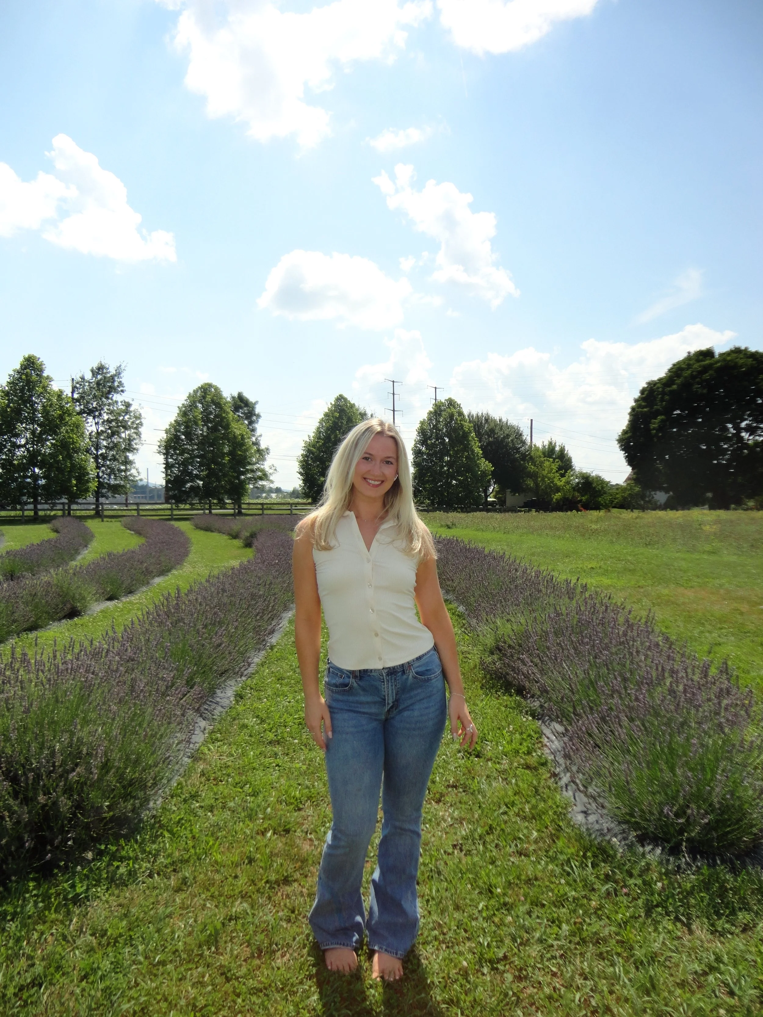 A woman with long blonde hair smiling, standing in a field of lavender with rows of purple plants, green grass, and trees under a bright blue sky with scattered white clouds.
