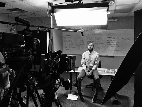 A man sitting on a chair in a recording or filming studio, with professional camera equipment and a large softbox lighting setup. A whiteboard with writing is visible in the background.